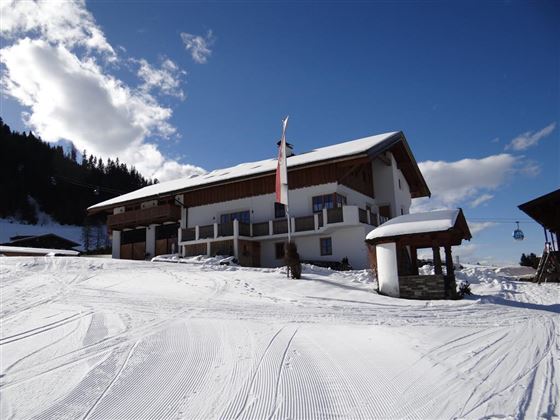Ein charmantes Berghaus in einer verschneiten Landschaft. Der Himmel ist blau mit wenigen Wolken und die Piste ist gut präpariert.