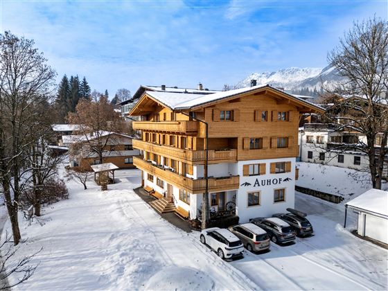 Ein gemütliches Holzgebäude im Schnee, umgeben von Bäumen. Es gibt mehrere Autos im Vordergrund und eine Berglandschaft im Hintergrund.