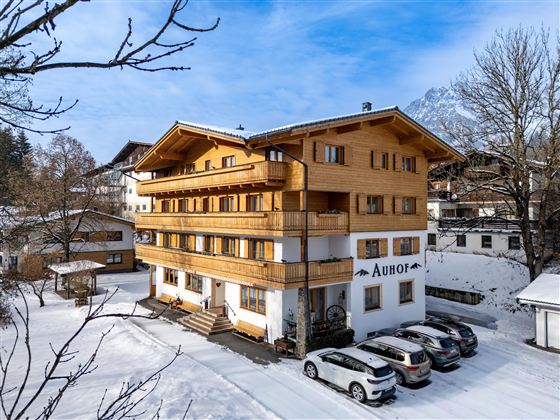 Ein modernes Holzgebäude in einer schneebedeckten Landschaft. Vor dem Gebäude stehen mehrere Autos und der Himmel ist klar.