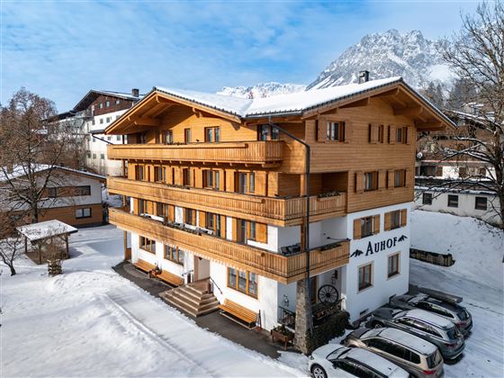 Ein charmantes Holzhaus im Schnee mit mehreren Etagen und Balkonen. Im Hintergrund sind schneebedeckte Berge und ein klarer Himmel sichtbar.