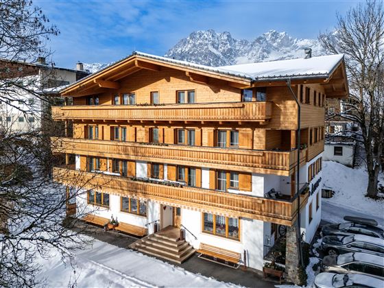 Ein gemütliches Holzhaus im Schnee mit mehreren Balkonen. Im Hintergrund sind majestätische Berge und ein klarer blauer Himmel zu sehen.