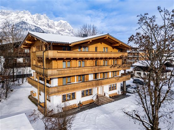 Ein gemütliches Holzgebäude im Schnee, umgeben von Baumkronen. Im Hintergrund sind majestätische Berge sichtbar.