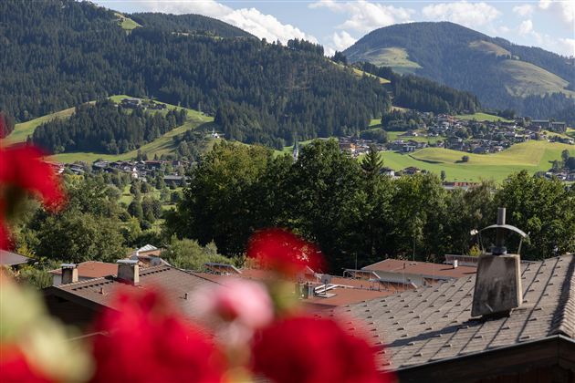 Eine malerische Berglandschaft mit grünen Hügeln und einem klaren Himmel. Im Vordergrund sind blühende Blumen und Dächer von Häusern zu sehen.<br/>Zeigt den Ausblick auf Ellmau vom Balkon aus.