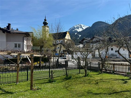 An idyllic village with beautiful houses and a church tower. In the background, snow-covered mountains and a clear blue sky can be seen.