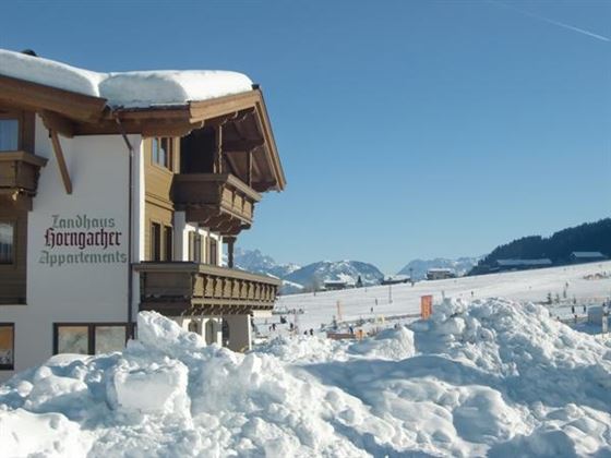 Ein charmantes Ferienhaus im Schnee mit traditionellen Architekturelementen. Im Hintergrund erstrecken sich die schneebedeckten Berge unter einem strahlend blauen Himmel.