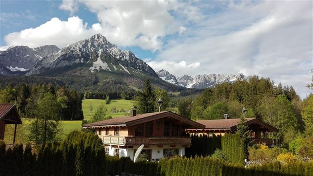 Eine idyllische Berglandschaft mit schneebedeckten Gipfeln und sattgrünen Wiesen. Im Vordergrund steht ein gemütliches Holzhaus umgeben von Bäumen.