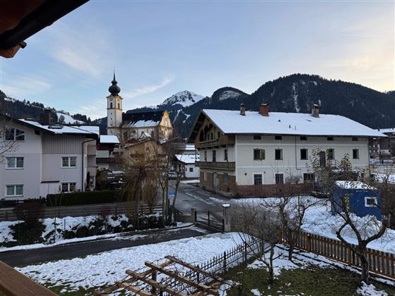 Eine malerische Winterlandschaft mit schneebedeckten Dächern. Im Hintergrund sind Berge und eine Kirche zu sehen.
