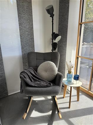 Een gezellige stoel met een kussen en een deken staat naast een kleine tafel. Op de achtergrond zijn een vloerlamp en grote ramen zichtbaar.