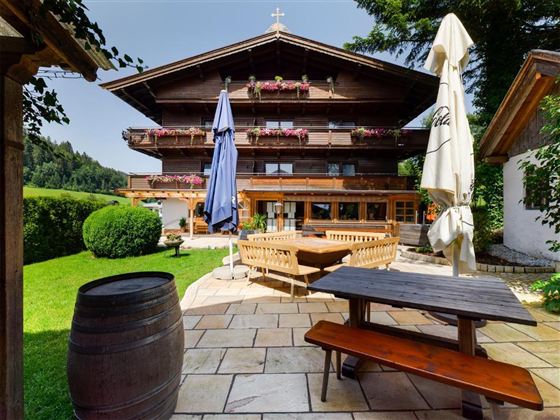 A traditional wooden house with blooming balcony boxes, surrounded by a beautiful green landscape. In the foreground are wooden furniture and a large sunshade.