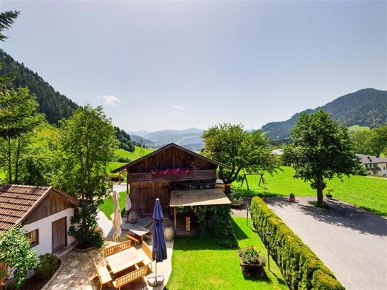 A picturesque landscape with a traditional wooden house and green meadows. In the background, gentle mountains and a clear sky can be seen.