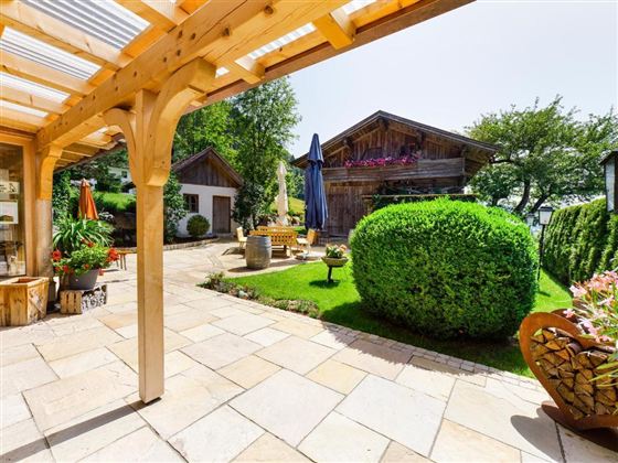 An inviting garden with neatly trimmed hedges and blooming plants. In the background, traditional wooden houses and a sunny terrace can be seen.