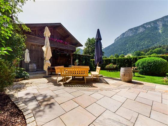 A beautiful garden with a stone terrace and cozy seating. In the background, mountains and lush scenery can be seen.