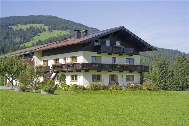 Ein charmantes Landhaus mit einem großen Garten und Balkonen. Im Hintergrund erstrecken sich grüne Hügel und Bäume.