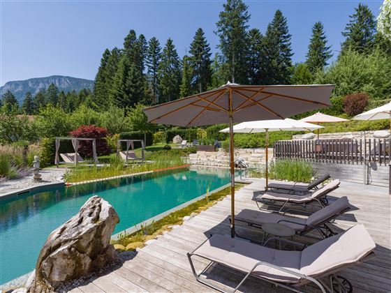 Ruhiger Gartenbereich des Hotel Alpenpanorama mit Sonnenliegen, Sonnenschirm und Naturpool im Hintergrund.