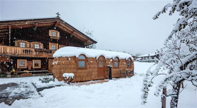 A traditional wooden house with a snow-covered roof sits in a wintry landscape. Snow blankets the ground and the trees all around.