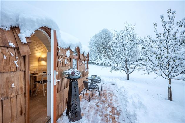 A cozy wooden house in the snow with a snowy landscape. The entrance is inviting, with a table and chairs outside.