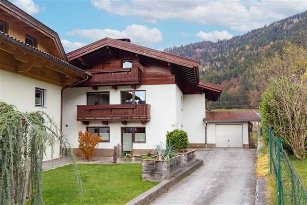 Ein gemütliches Haus im alpinen Stil mit einem grünen Garten. Im Hintergrund sind sanfte Hügel und Bäume zu sehen.