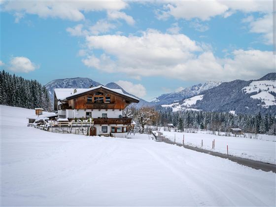A picturesque mountain landscape in winter with snow-covered fields. In the foreground, there is a traditional wooden house against an impressive mountain backdrop.