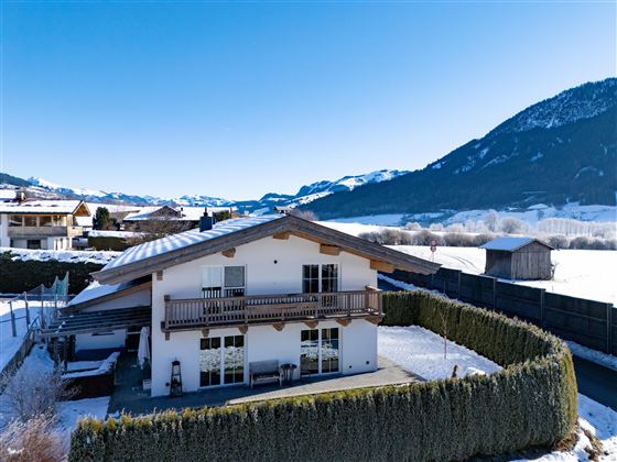 Ein gemütliches Haus im Winter mit einer schneebedeckten Landschaft. Im Hintergrund sind Berge und ein klarer blauer Himmel zu sehen.