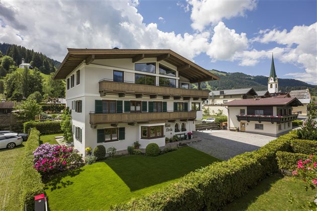 A modern building surrounded by green spaces and flowers. In the background, a church tower and the mountains can be seen.
