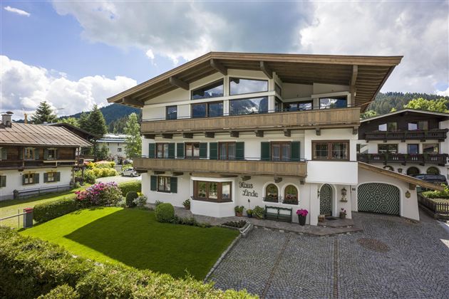 A modern house with a large balcony and a beautiful garden landscape. The surroundings are green and surrounded by mountains.