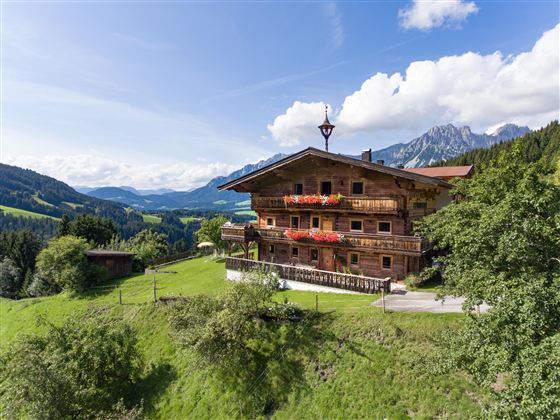 Een mooi houten huisje in het berglandschap.  
Omringd door groene weilanden en majestueuze bergen onder een blauwe hemel.