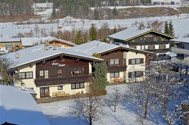Ein traditionelles Bauernhaus umgeben von schneebedeckten Bergen und Bäumen. Der Himmel ist klar und blau, was eine ruhige Atmosphäre schafft.