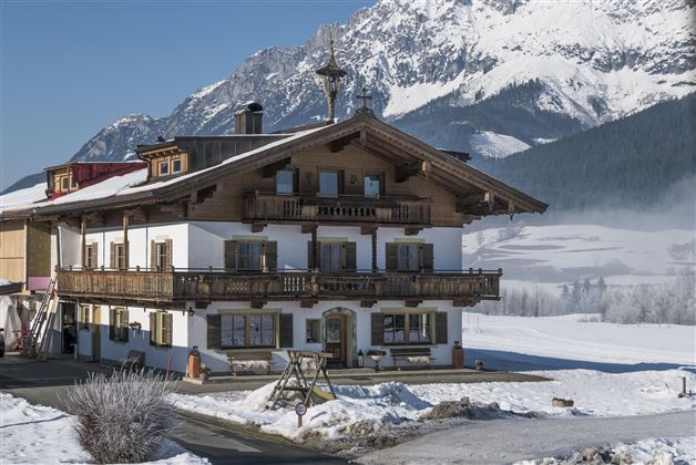 Ein traditionelles Alpenhaus im Schnee mit umliegenden Bergen. Die sonnige Landschaft vermittelt eine ruhige Winteratmosphäre.