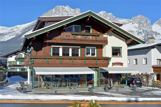 Ein traditionelles Alpenhaus mit Holzverkleidung und Balkon. Im Vordergrund sind schneebedeckte Bereiche und die beeindruckenden Berge im Hintergrund sichtbar.