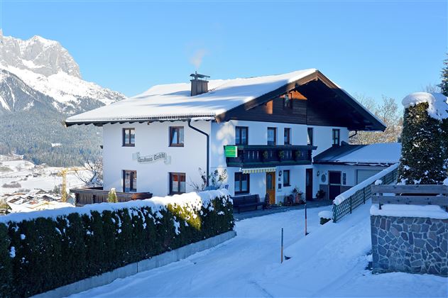 A picturesque house in the snow with a beautiful mountain landscape in the background. The sky is clear and blue, creating a tranquil winter atmosphere.