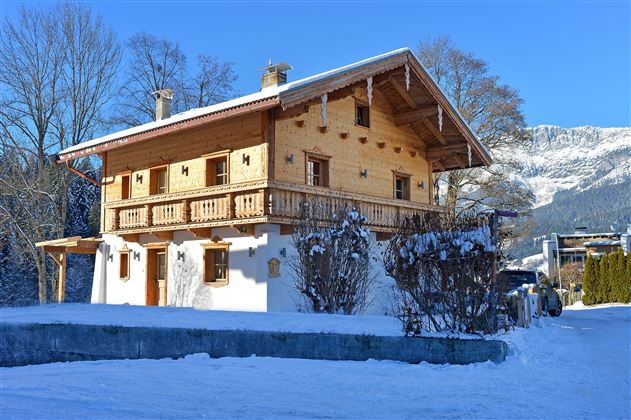 Ein traditionelles Holzhaus in einer schneebedeckten Landschaft. Im Hintergrund sind Bäume und Berge zu sehen.