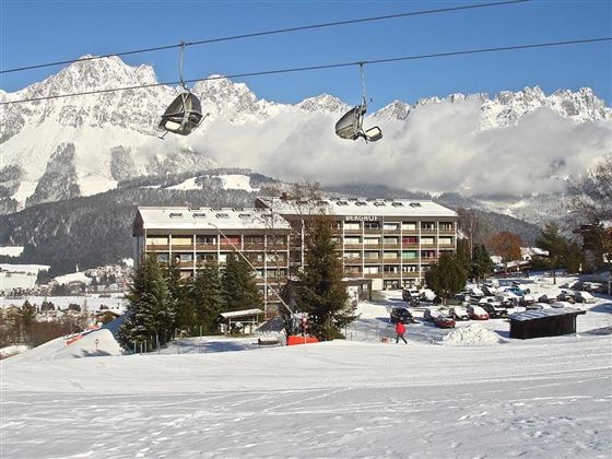 Ein schneebedeckter Winterort mit einem Gebäude und einer Seilbahn im Vordergrund. Im Hintergrund sind hohe Berge und ein klarer Himmel zu sehen.