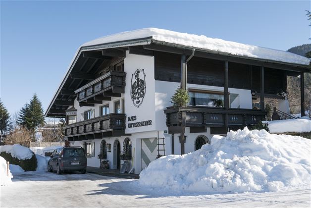Ein traditionelles Haus im Alpenstil, umgeben von Schnee. Die Fassade ist weiß mit dunklen Balkonen und einer stilvollen Beschriftung.