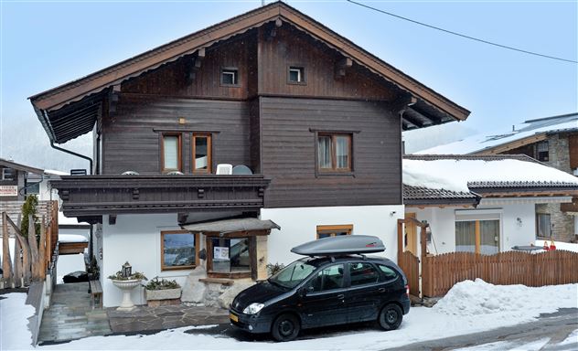 Ein traditionelles Holzhaus im Schnee mit einem schwarzen Auto davor. Die Umgebung ist ruhiger und winterlich.