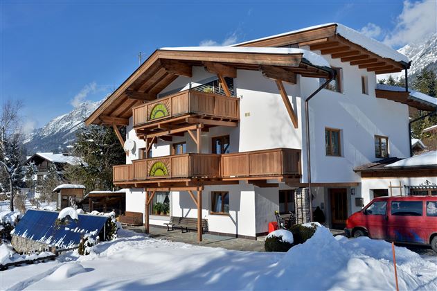 A beautiful old house with wooden balustrades, surrounded by snow. In the background, mountains and blue sky can be seen.