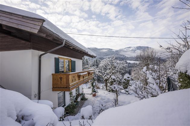 Ein charmantes Haus im Winter, umgeben von schneebedeckten Bäumen und Hügeln. Der Himmel ist klar mit einigen Wolken.