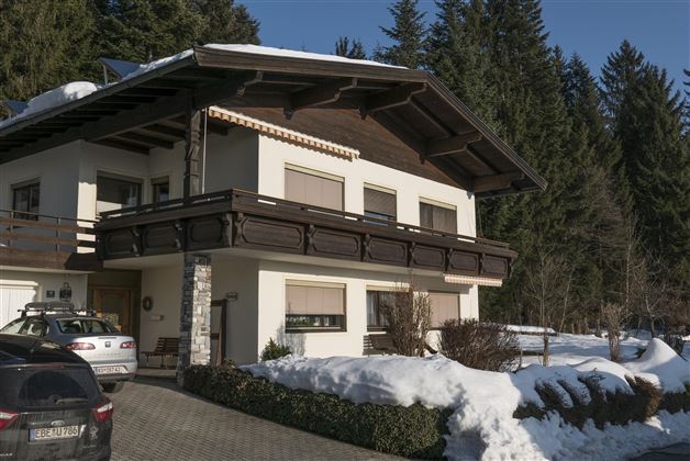 A modern house in alpine style with a balcony and a snow-covered surroundings. In the background, green trees can be seen.