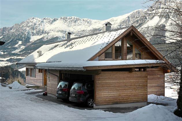 A modern wooden house in the snow, surrounded by mountains. Two cars are parked in the garage.