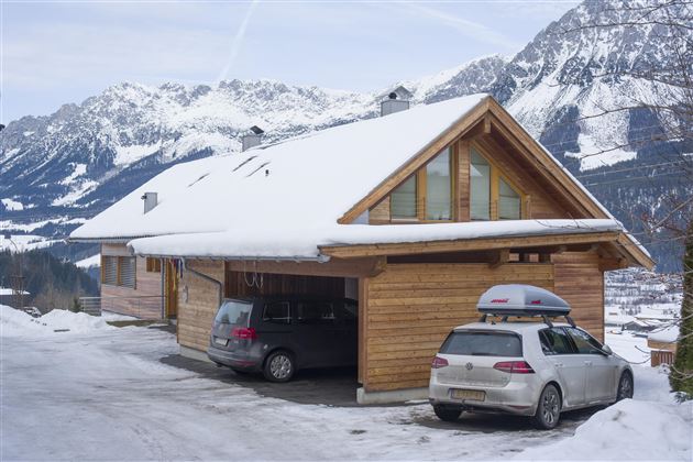 A cozy wooden house in the snow with a garage area. Impressive mountains can be seen in the background.