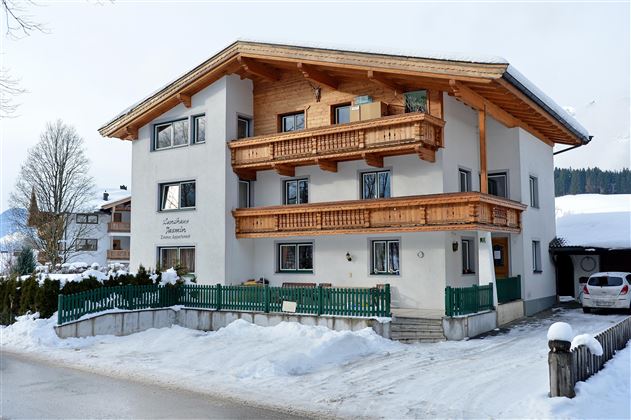 A three-story building in alpine style with wooden cladding and balconies. It is situated in a snowy environment, surrounded by trees and other buildings.