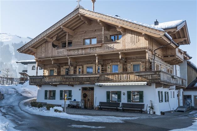 A traditional wooden house with several balconies and large windows. It stands in a snow-covered landscape and seems to be nestled in a picturesque mountain setting.