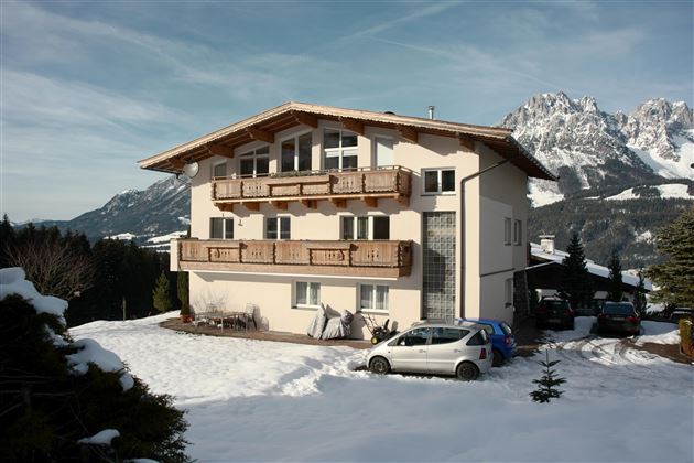 Ein großes, modernes Haus im Schnee mit einem Balkon und Sicht auf die Berge. Um das Haus parken einige Autos.