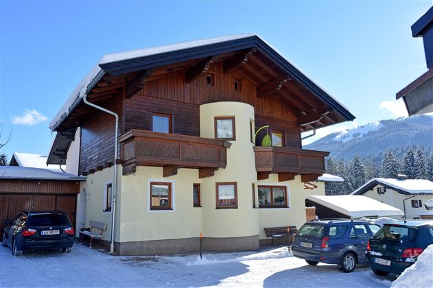 Ein zweigeschossiges Haus mit einem holzverkleideten oberen Stockwerk. Es liegt in einer schneebedeckten Umgebung und hat einen klaren blauen Himmel im Hintergrund.