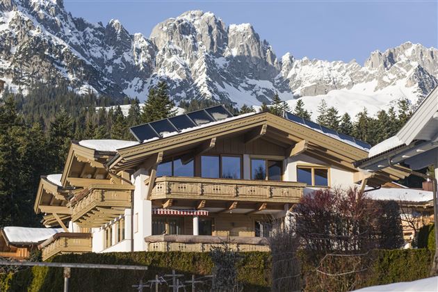 Ein modernes Chalet in den Bergen mit schneebedeckten Gipfeln im Hintergrund. Die Architektur kombiniert Holz und große Fenster, die viel Licht bringen.