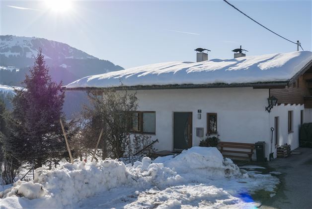 A cozy house in the snow with a snow-covered roof. In the background, there are mountains and a clear blue sky.