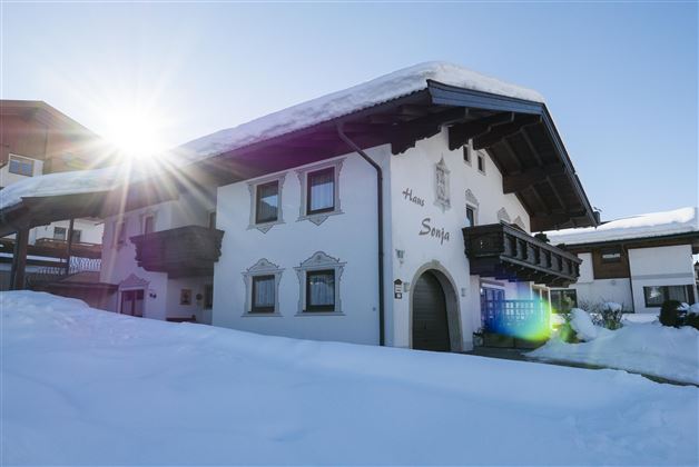 Ein charmantes, traditionelles Haus im Schnee, mit einem klaren blauen Himmel im Hintergrund. Die Sonne strahlt in das Bild und erzeugt eine warme Atmosphäre.