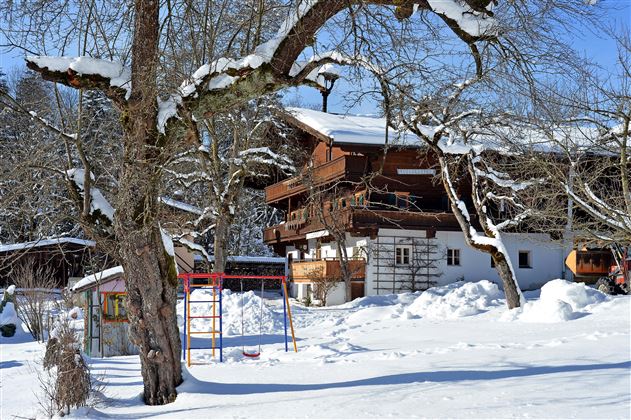 Ein schneebedecktes Haus umgeben von Bäumen. Im Vordergrund ist ein Spielplatz mit einer Schaukel zu sehen.