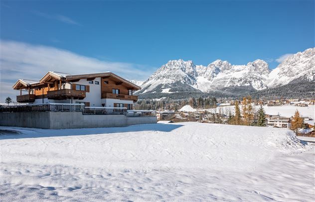 Ein modernes Holzhaus in einer verschneiten Landschaft. Im Hintergrund ragen majestätische Berge unter einem klaren blauen Himmel empor.