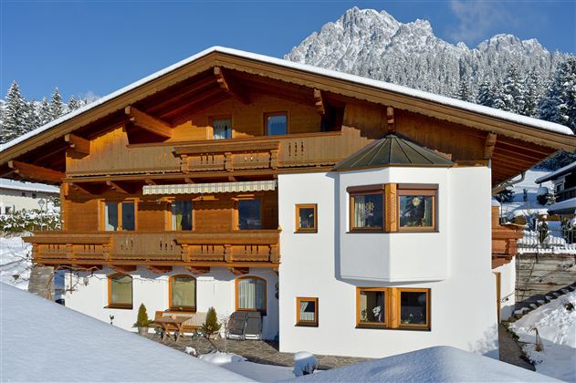 A chalet-shaped wooden house with several balconies stands in the snow. Snow-covered mountains can be seen in the background.