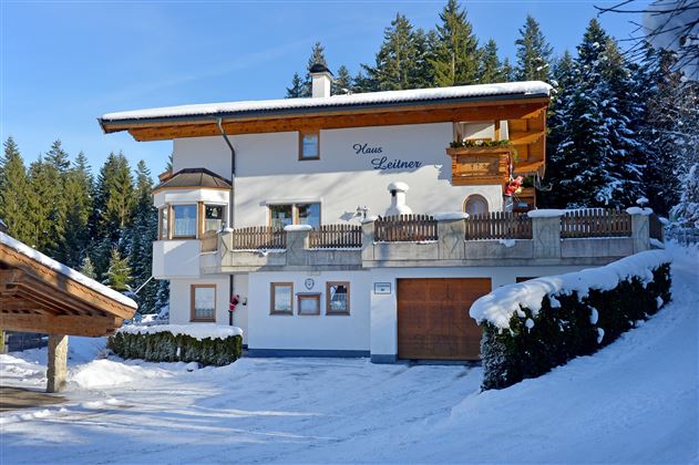 Ein hübsches, weißes Haus im Schnee, umgeben von Tannenbäumen. Der Balkon hat eine schöne Holzverkleidung und es gibt eine Garage.